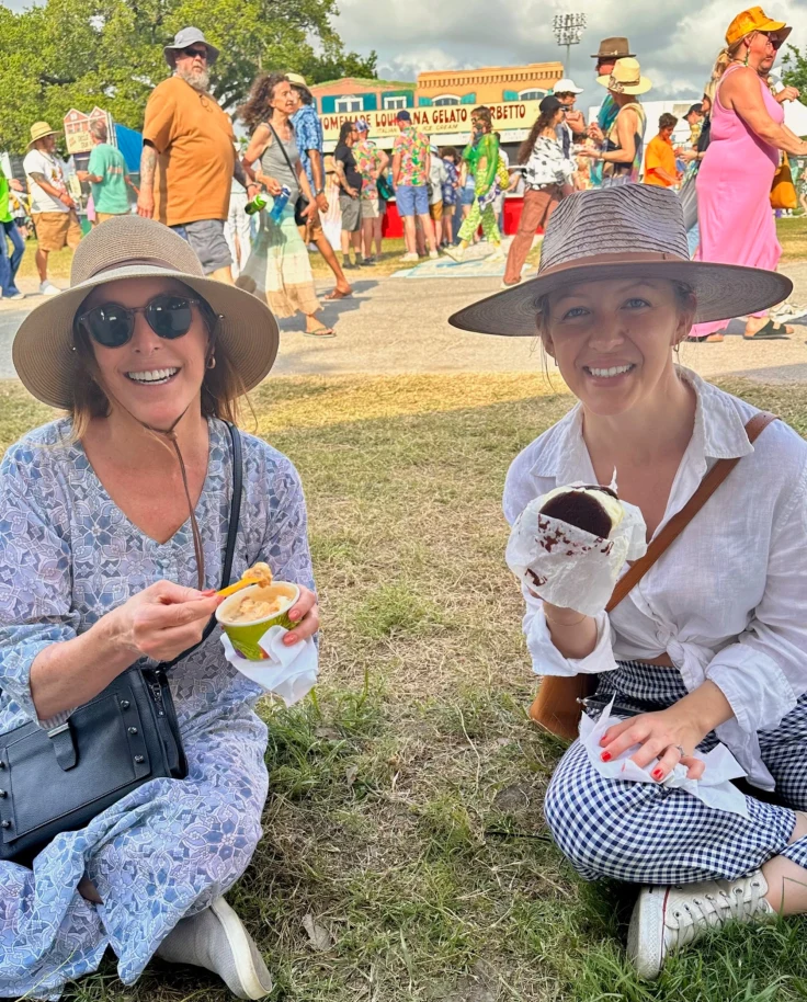 Allison and a friend in straw hats sitting on a lawn eating ice cream, with a lively crowd and food stalls in the background.