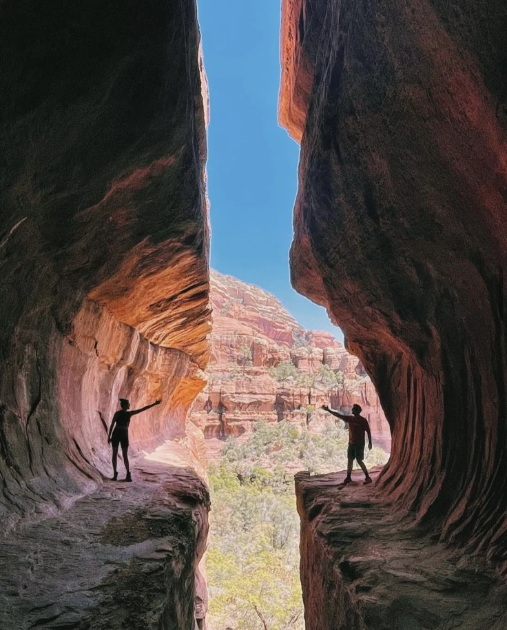 Two silhouettes stand at the edge of a natural divide, bridging the gap between towering cliffs under a vast blue sky.