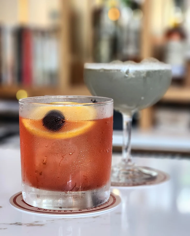 The image shows a chilled beverage with ice, garnished with an orange slice and a dark-colored berry, set on a coaster.