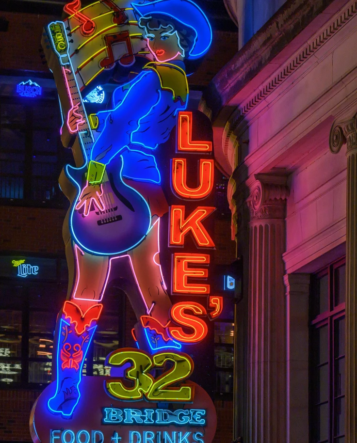 A neon sign with a woman wearing a cowboy hat and holding a guitar that reads "Lukes" at night.