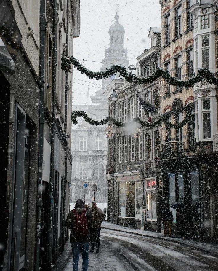 View of a narrow street with few pedestrians in light snow decorated with Christmas garlands above