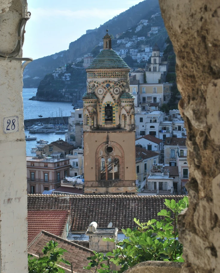 A stone archway highlighting an old bell tower and coastal town in the distance with the sea and mountains showing.