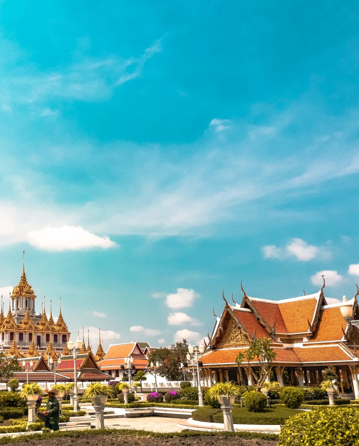 Temples in Bangkok, Thailand with a blue cloudy sky, green groomed bushes, and white paths with people walking on them.