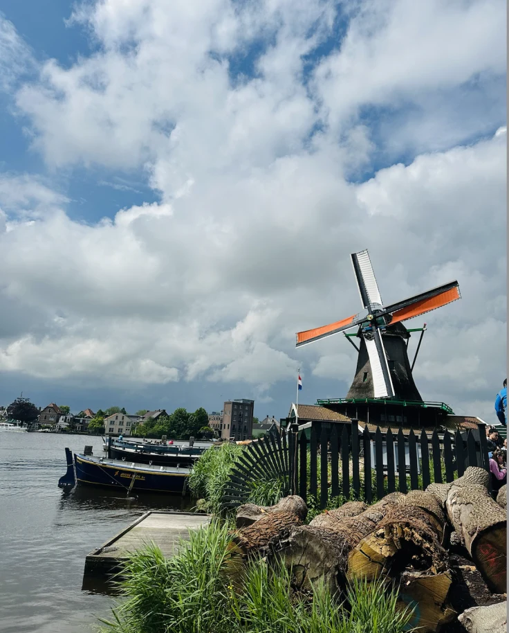 A windmill standing on the edge of a body of water with canoes parked next to it and buildings in the distance along the shore line.