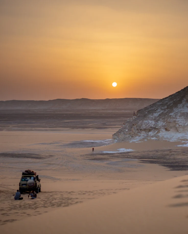sunset over a calm desert with a car and a person walking towards the sun