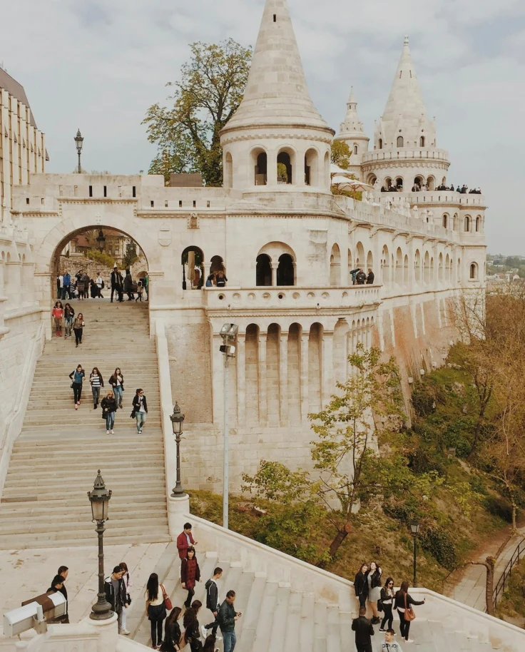 A picture of an old stone building with detailed architecture, steps, towers and trees. There are people walking down the steps and in front of the building.