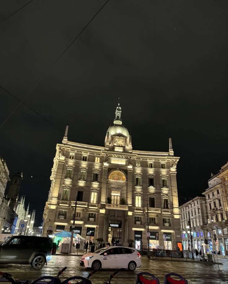 A low-angled shot of a beige-colored building near a street at night. There are cars driving in front of it.