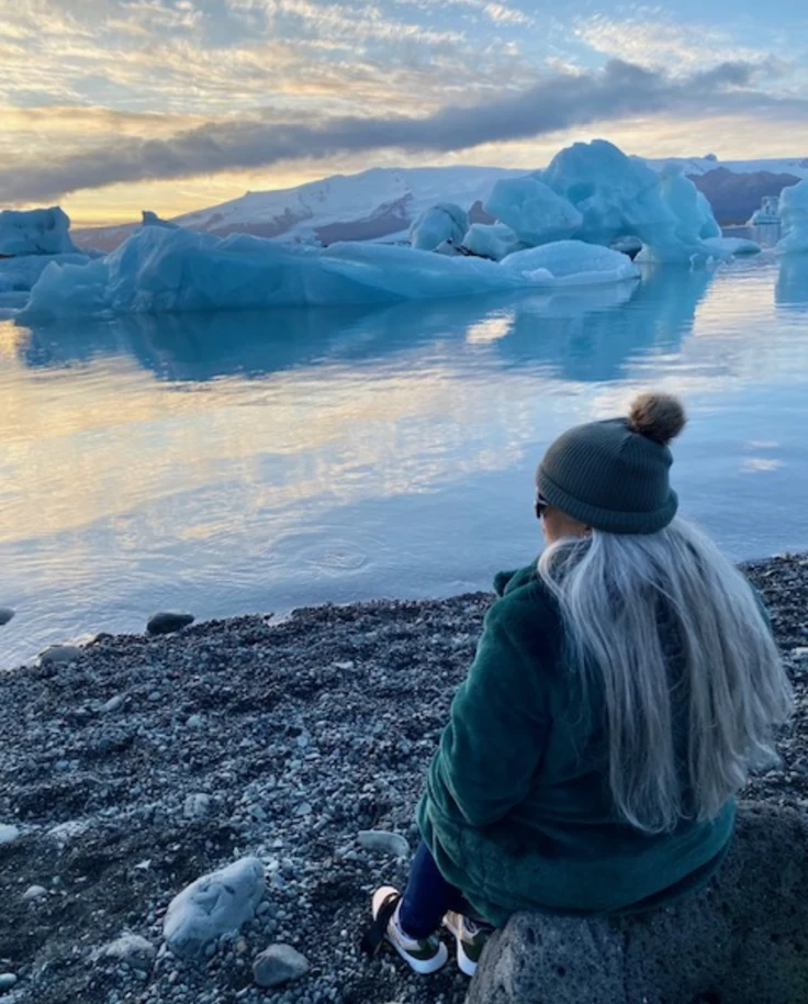 This image depicts a blonde woman sitting on a rocky shore and looking out onto a clear lake with ice bergs in the surrounding areas.