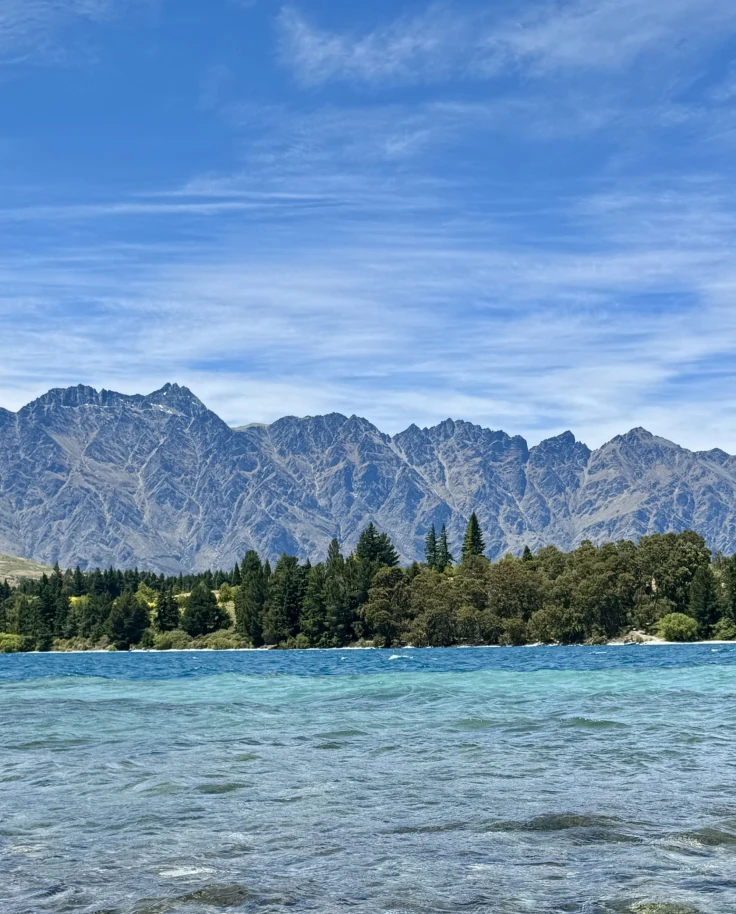 A vibrant landscape with a clear blue lake in the foreground and rugged mountains in the background under a partly cloudy sky.