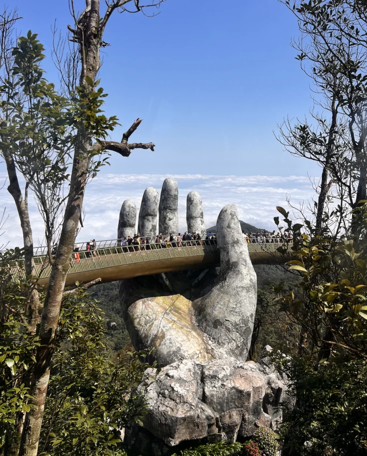 A view of a large stone hand with a bridge built onto it and people walking across. There are also two sets of tall trees and a forest in the surrounding areas.
