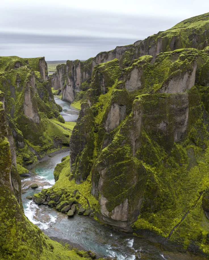 Green moss on tall, large gray rocky mountains with a gushing river flowing through the center on an Iceland itinerary.