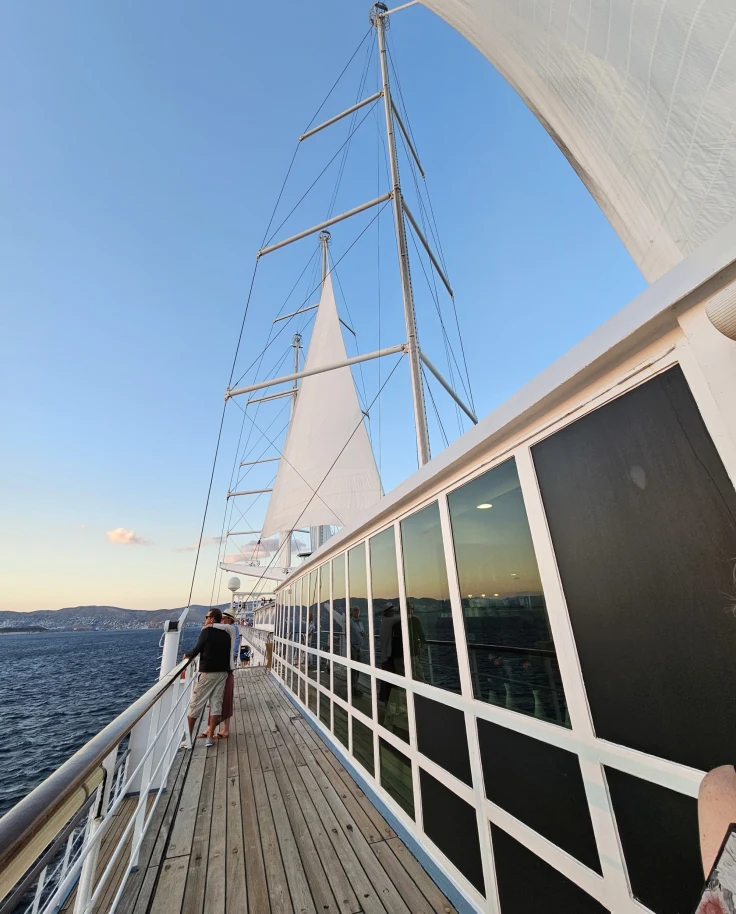 The image depicts a sunset view from the deck of a sailboat with sails unfurled and the horizon in the background.