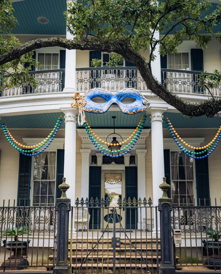 The image depicts a two-story house adorned with Mardi Gras beads and a large blue mask