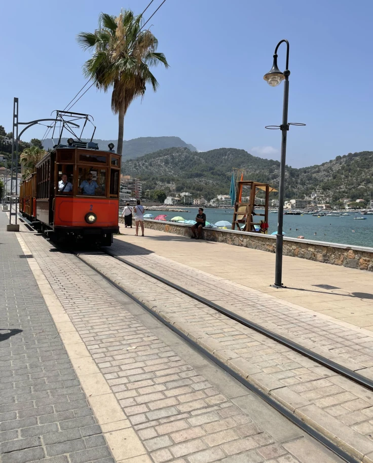 A red tram car chugging down a coastal road with the sea, mountains and blue sky in the background.