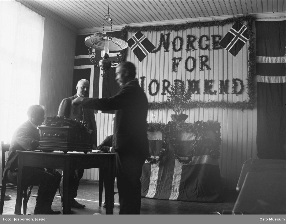 Foto fra valglokale i 1905. En mann putter stemmen sin i urnen mens to funksjonærer ser på. På veggen bak henger store flagg, kranser av løv og mottoet «Norge for nordmenn» med store bokstaver.