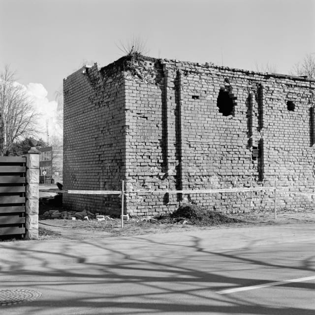 The corner of an old brick building, with a tape blocking access to it. In the foreground, the branches of a tree lead up to the house. In the distance, a bright cloud.