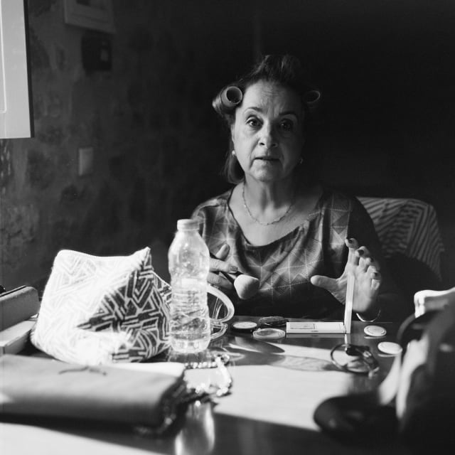 Mom sat at a table indoors, putting on makeup. The table seems neater, and a water bottle still holds the mirror. She is holding a brush, and looking directly at the camera, looking about to speak.