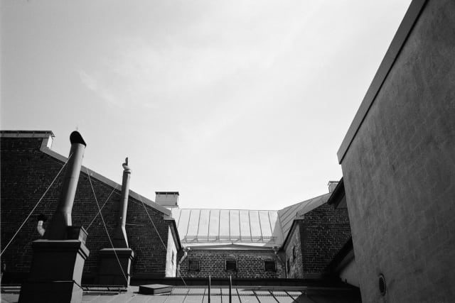 A view upwards onto rooftops. Chimneys, maintenance ladders, and ventilation shafts line the tops. The light is coming from a right angle beyond the frame, casting hard shadows where they appear.