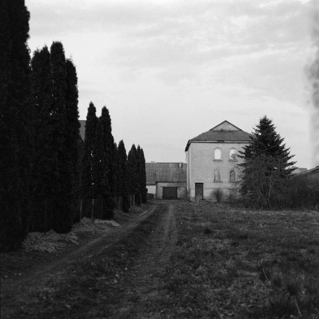 A two story house in the distance, with a long garden or field leading up to it. On the left, a row of tall dark trees. On the right, a smudge or smoke. The house is lit by a distant light.