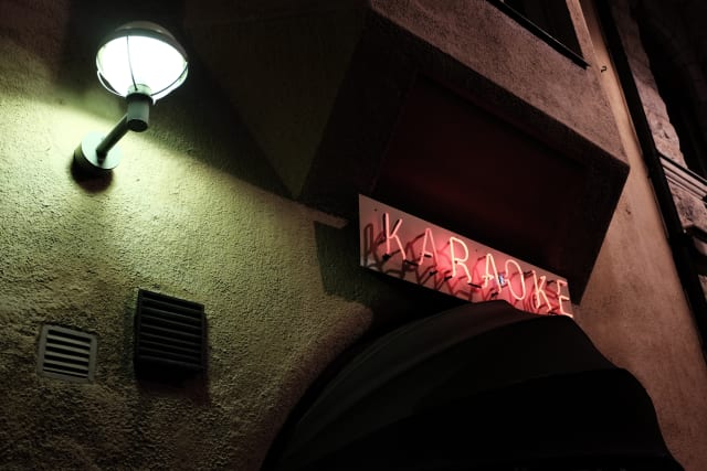 The red neon sign of a Karaoke bar, by the pale green glow of a street lamp.