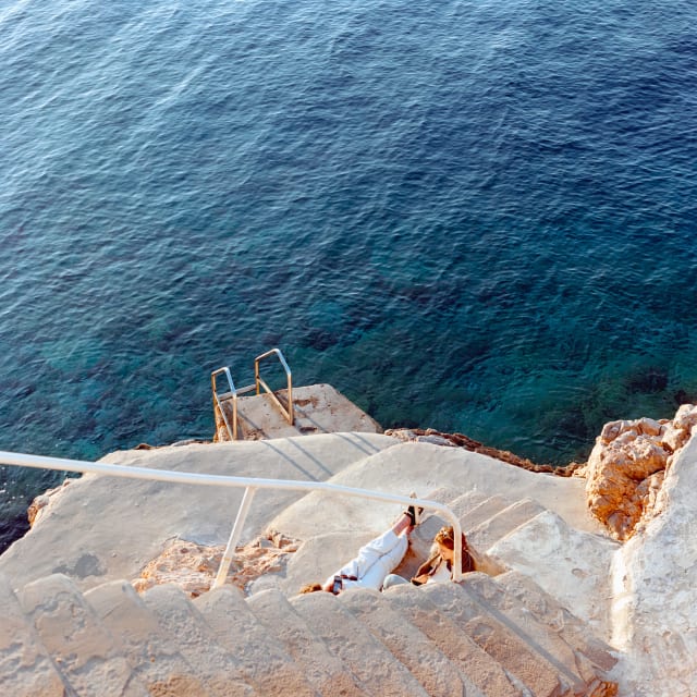 A view down stone steps toward the sea. Two people are lying down to the side of the steps, reading. The scene is filled with sunset light, off the frame. A deep blue sea covers the top two thirds.