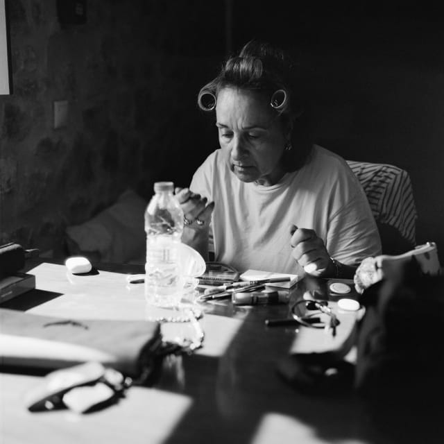 Mom sat at a table indoors, putting on makeup. The table has a random assortment of things, from makeup utensils, to a purse, and a water bottle holding up a mirror. The photo is contrasty, with some bright light shining from a window on the left.