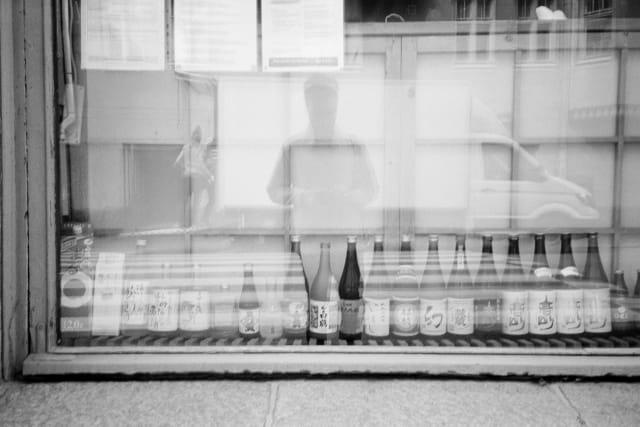 A shop window, lined with bottles. The photographer is reflected with their hands on the camera a bit above waist level. A truck with people loading it is reflected behind them.