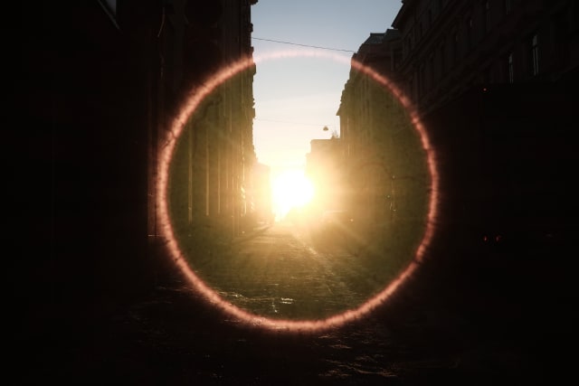 A mostly shaded city on the sides and bottom. The sun is intensely shining in the middle, with a bright red halo that almost sears the image.