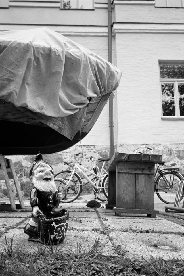 A garden gnome viewed from the ground level, with a skull and bones bucket in front of them. They have a smirk and some cool lipstick. Behind them, there are bikes, a small boat under a tarp, and an old building.