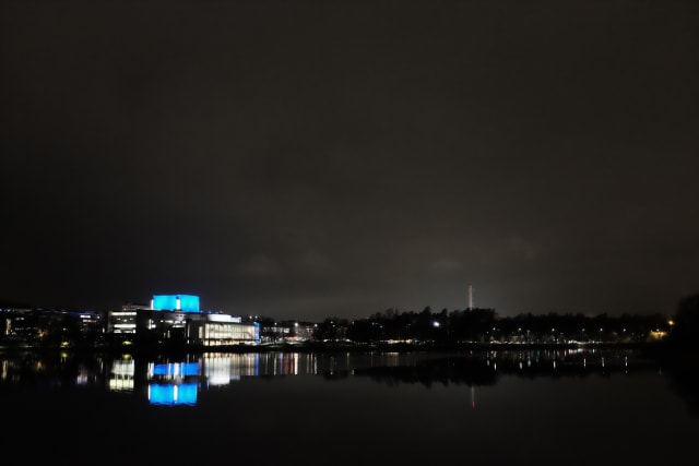 A white and blue building in the distance, over water. The building and trees are reflected on the water's surface.