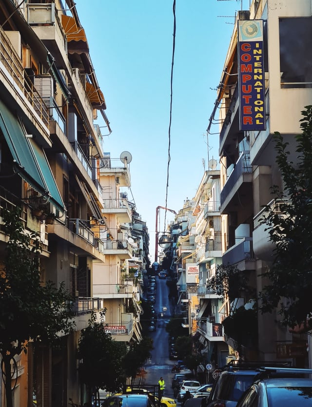 A city street, with multi-storey buildings on each side. The street has a steep incline in the distance, with cars, electricity cables, and advertising signs lining the sky towards it.