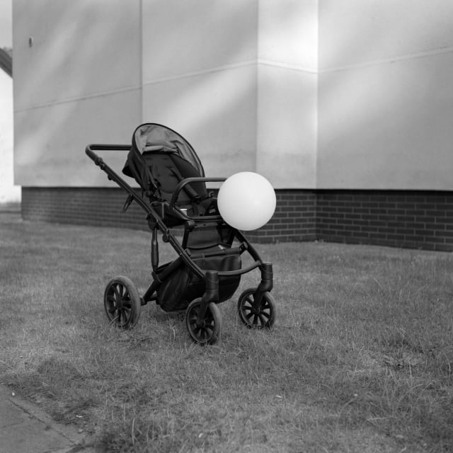 A baby stroller stands on a patch of grass, in front of a softly-lit building. The stroller is empty, save for a balloon tied to its front. The balloon seems to be drooping slightly.