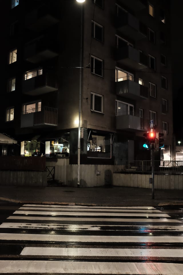 A street corner, with a pedestrian crossing leading up to it. Surfaces reflect street lights after the rain. A person is sitting at a bar, while above it apartment lights are on.