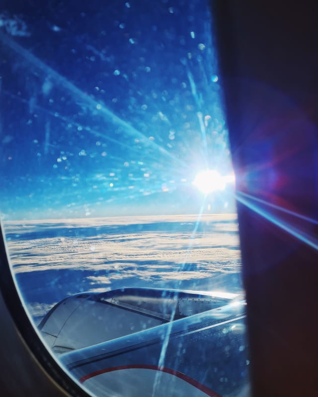 An airplane window. The sun, or maybe a reflection of it, shines prominently over a sea of clouds, flaring across the frame.