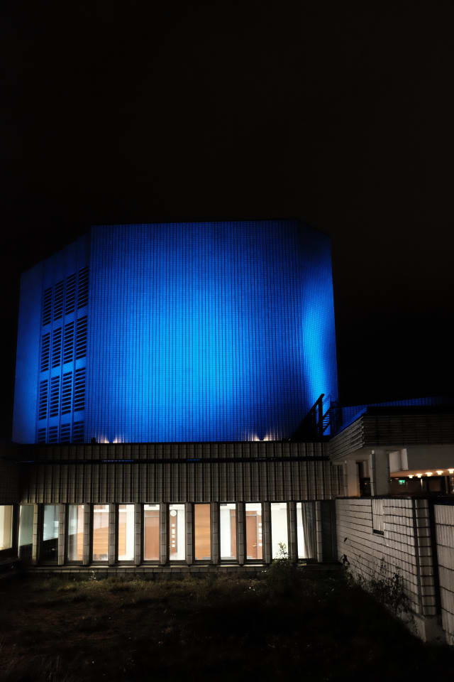 A blue, tiled building nearby. Tiles surround a small garden, and bright lights are on inside.