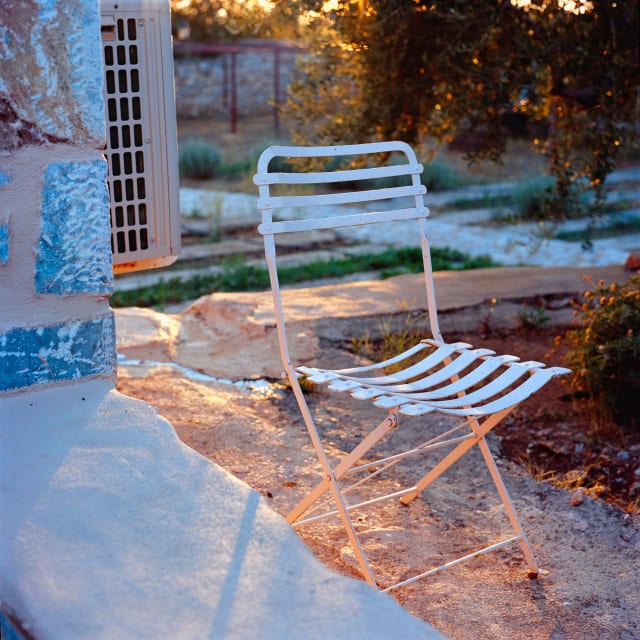 A square frame with a white metal chair with grills standing in the middle. Its seat and back trace a similar shape as the stone porch on the foreground. The sun casts rays down the middle of the frame, and small shadows from the chair. There is a warmth and stillness in the scene.