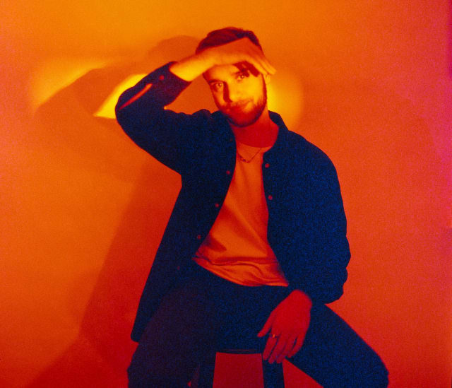 Myself sitting on a chair or stool, looking at the camera. There is a soft smile on my face. The room is filled with a red-orange light, and a brighter line of light crosses my face. I am lifting my hand to shield my left eye from the light.
