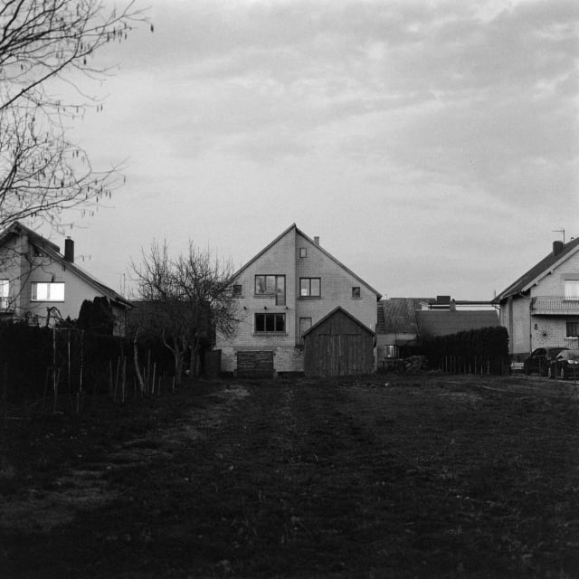 A two story house in the distance, with a garden or field leading up to it. The house itself is largely in the shade, but the ones adjacent to it have their windows lit up.