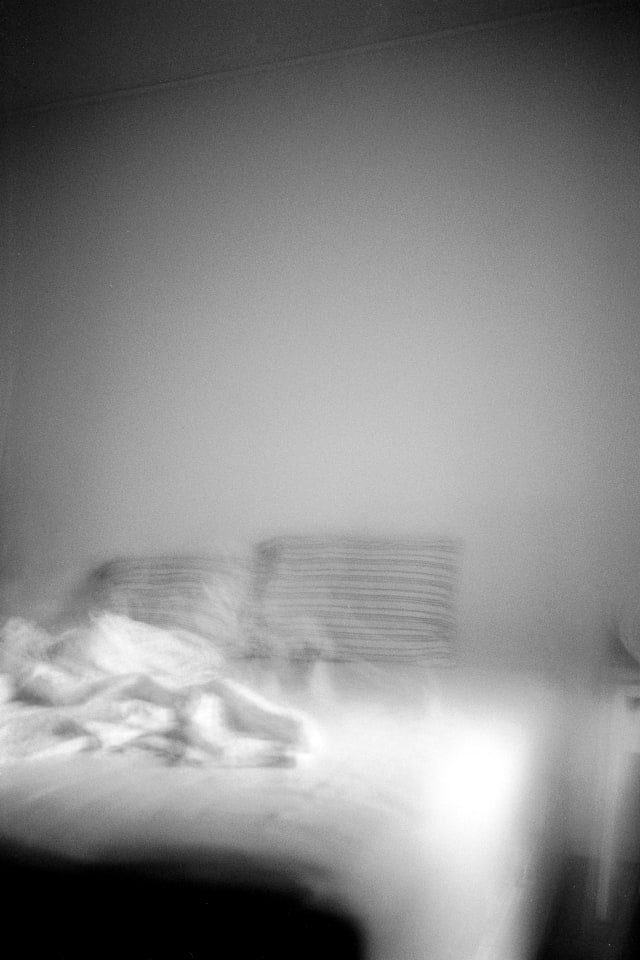 A blurry and shaky view of a bed or bedroom. Shadows line the edges of the frame. On the bed, two pillows are standing haphazardly, and a blanket is draped at an angle.