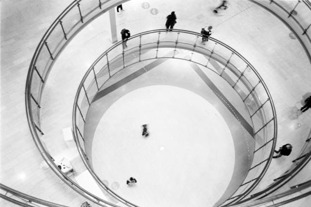 A top-down view of three floors of a shopping mall, through a circular opening. People are standing or walking around each floor. The circles of each floor are merging into each other, leading to a spiral-like effect.