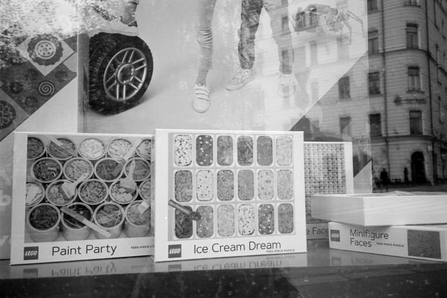 A shop window, with multiple reflections of the city on it. Boxes of LEGO line the display, titled "Paint Party", "Ice Cream Dream" and "Minifigure Faces".