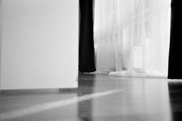 A wooden floor in the foreground, looking towards draped curtains in the back, at an angle. The light from the window is sharp and reaches the edge of the frame.