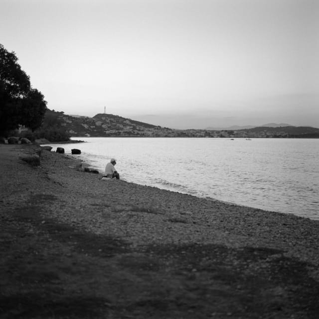 A person sitting by themself on a strip of beach after sunset. The beach leading up to them is darker, while the sea in front is lit, perhaps dimly, by the remainders of the sunlight. The person seems to be thinking and gazing. A tiny wave is about to crash at their feet.