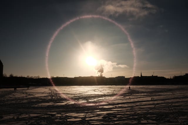 A landscape over Helsinki. The icy sea stretches over the bottom of the frame, leading to a shaded outline in the distance. The sun is in the middle casting a glare on the lens and landscape. Figures of people dot the ice.