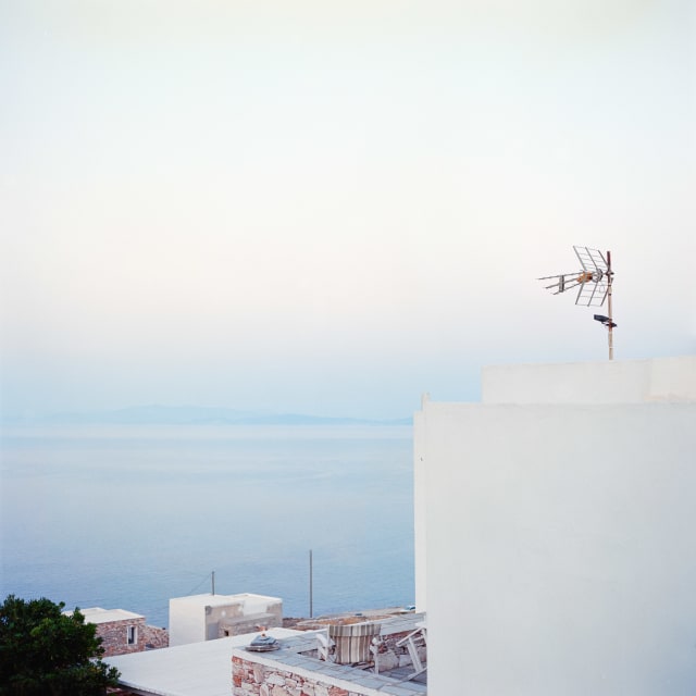 A square frame over white buildings and the sea. The dominant colours are a light blue and white. On a building on the right, a TV antenna is pointing towards the distance.