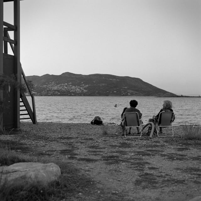 Two people sitting on portable chairs at the beach, near sunset. They seem to be talking with each other. On the left of the frame, a lifeguard post. In the distance, a blurry person is swimming. There is a visual path leading from the foreground of the frame to the water.