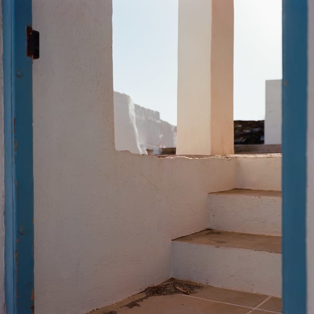 A view through a blue door frame, with no door to be found. A few steps are leading up to the camera level, and are lit by a warm light. In the distance, a white sky.