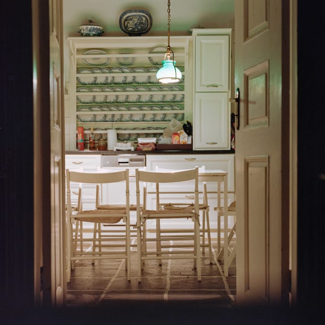 A view through two open white wooden doors, onto a kitchen. The kitchen is lit by a lamp hanging in the middle. The edges of the frame are pitch black.