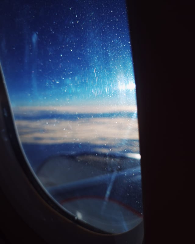 An airplane window, slightly blurry, with mostly the details and scratches on it being distinct. A reflection of the sun flares into rays near the edge of the window.