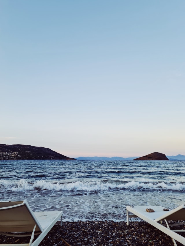 Two sunbeds by the sea, as a wave crashes. The sea is a deep blue, paired with a gradient in the sky.